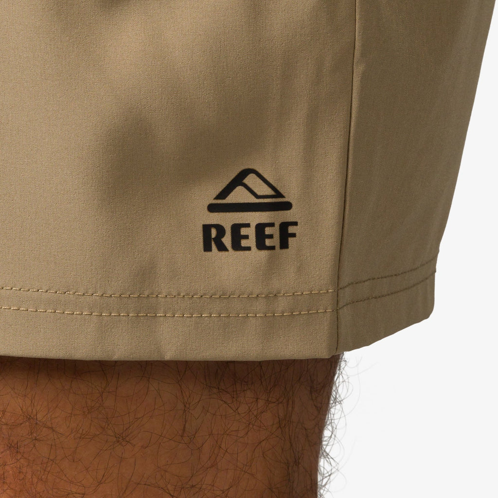Close-up of the bottom hem on the left leg of the Garland Hybrid Short in tan with black REEF logo, beach-inspired comfort.