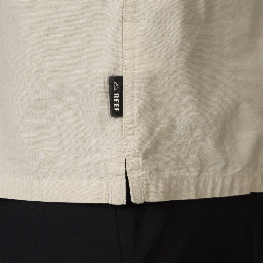 Close-up of the side hem detail on the Harold Camp Shirt showing REEF brand tag on beige, surf-inspired apparel.