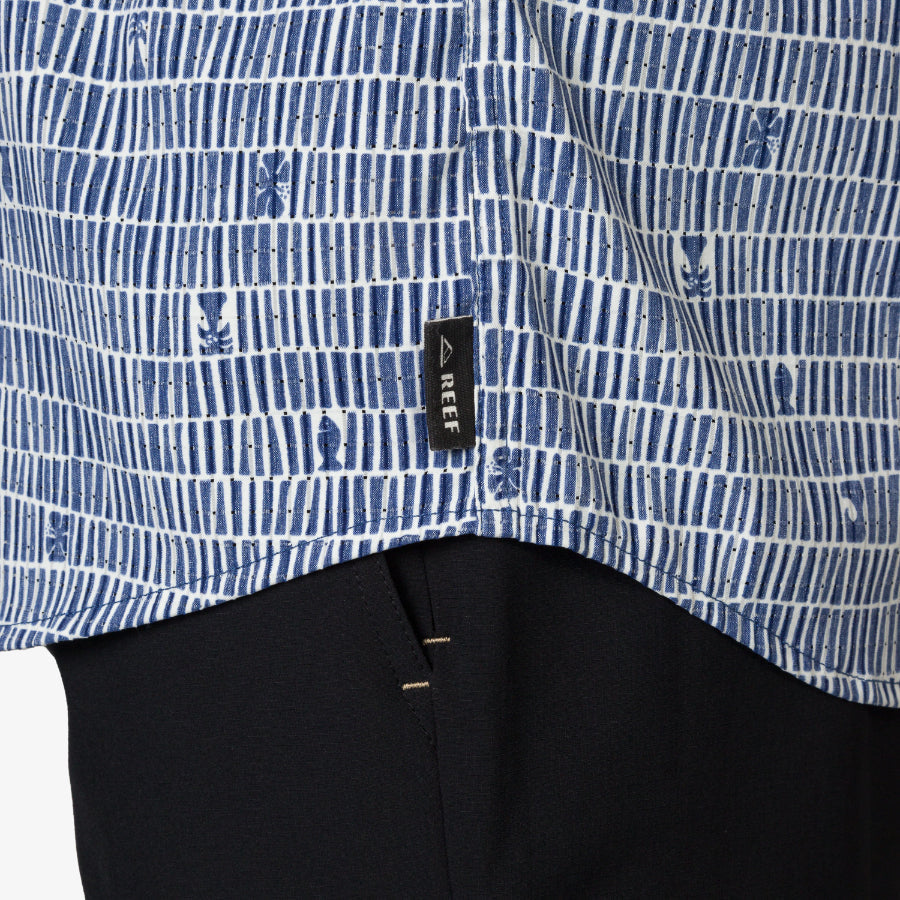 Close-up of blue patterned Ferris Classic Shirt hem with REEF tag over black pants, beach-inspired men's apparel.