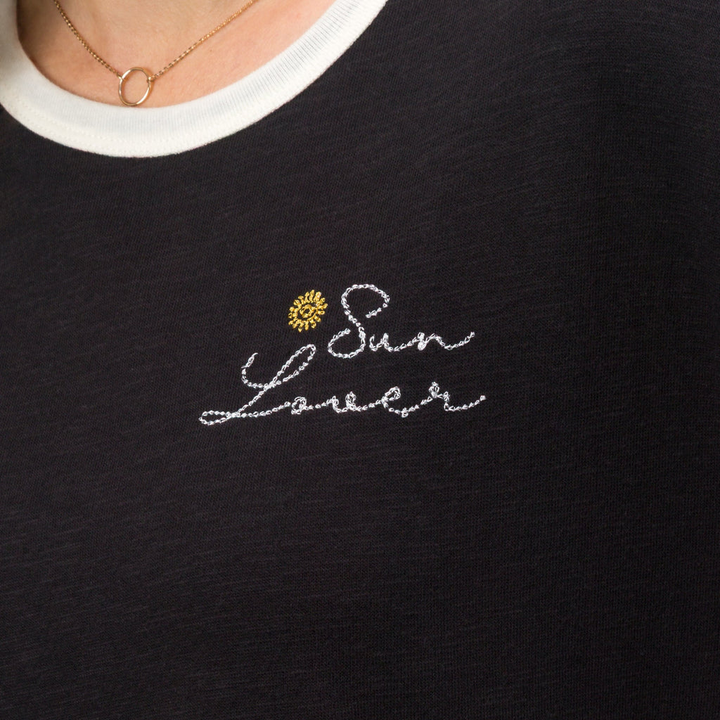 Close-up of the chest area on the Sun Lover Ringer Tee showing black fabric, white collar, and embroidered sun and text design.