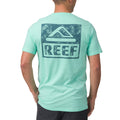 Man wearing light blue Wellie Too Tee facing away showing back logo, casual surf-inspired apparel for the beach.