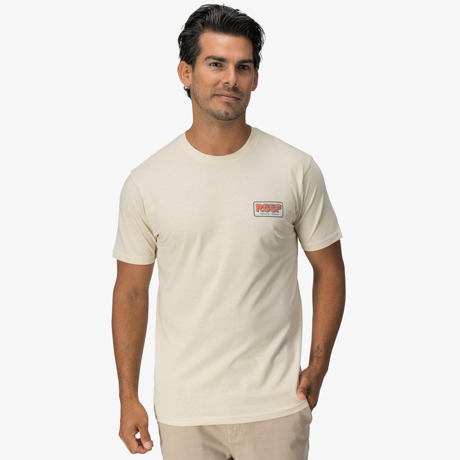 Man wearing a cream Quinn Tee with a small chest logo, surf-inspired, comfortable, and designed for beach lifestyles.