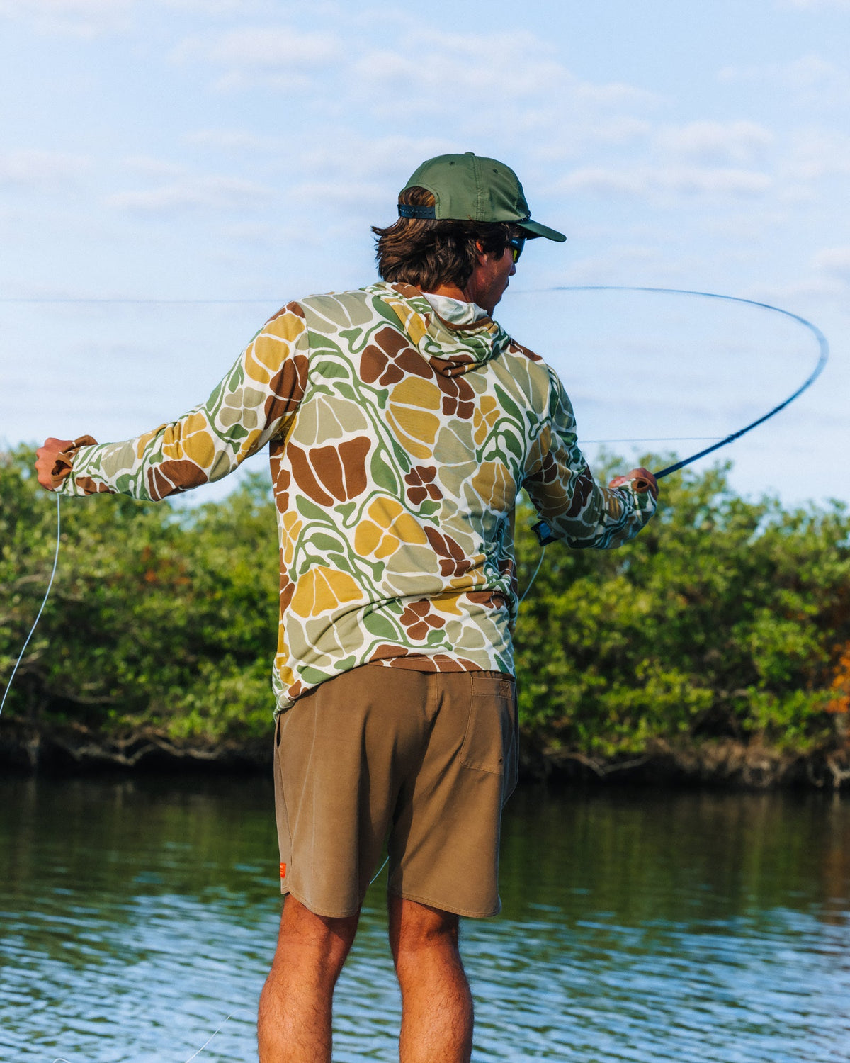 Man facing away wearing Men's Bamboo Lightweight Hoodie - Floral Camo casting fishing line by water on sunny day