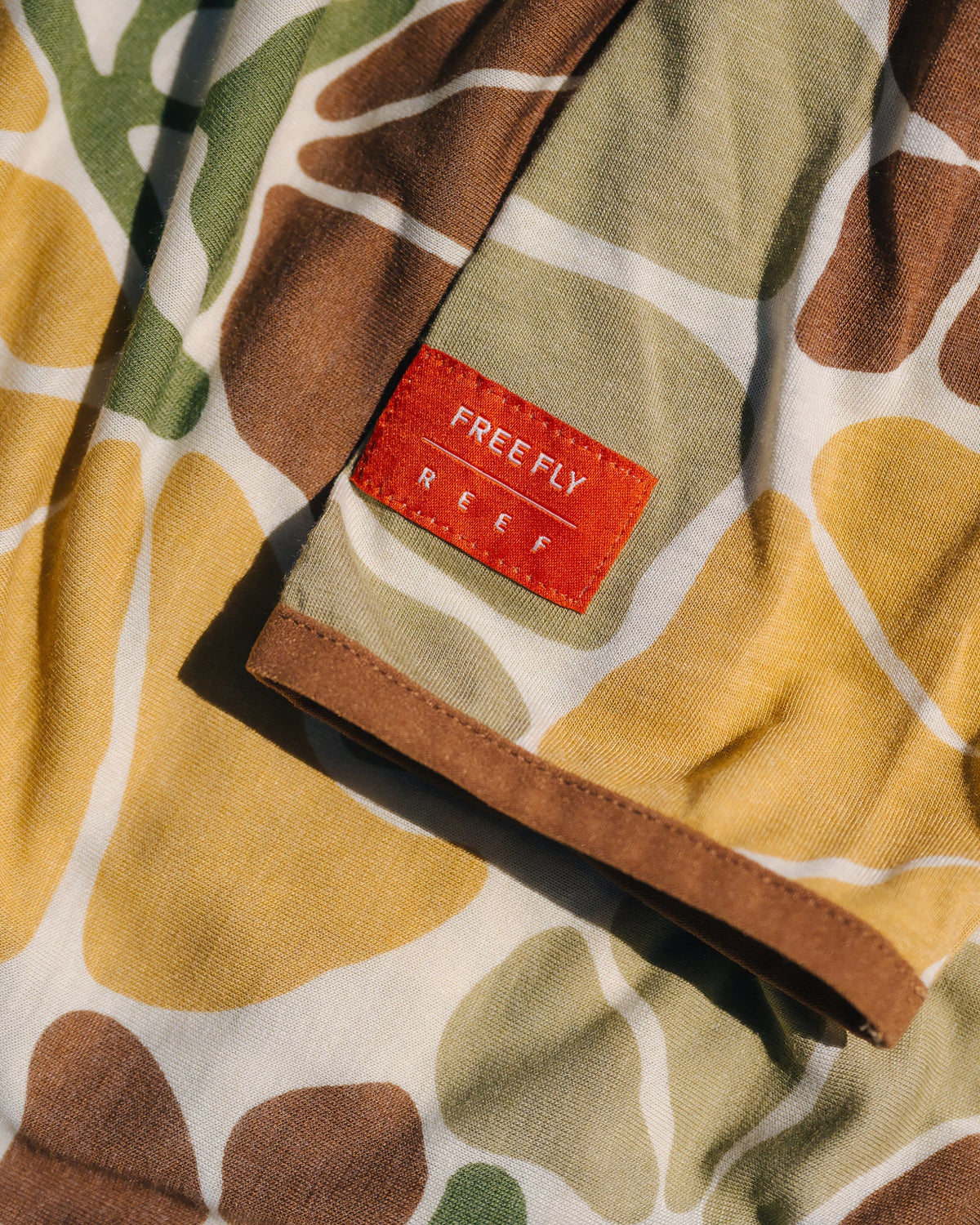 Close-up of Men's Bamboo Lightweight Hoodie - Floral Camo showing orange brand label, soft breathable fabric with earth tone camouflage design