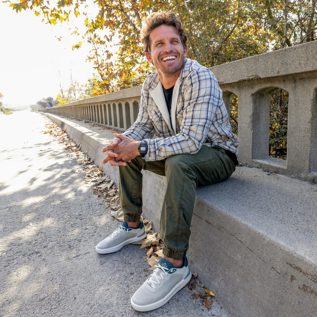 Man wearing Shoreline Del Mar sneakers facing forward outdoors sitting on concrete ledge with autumn leaves nearby