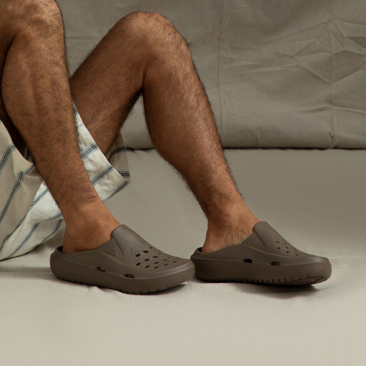 Pair of brown Glider slides facing left on man's feet, showing side and top in a close seated pose, beach-inspired comfort design.