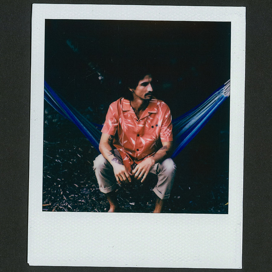 Man wearing a coral Kaden Camp Shirt sitting barefoot in front of a blue hammock, casual beach-inspired style.