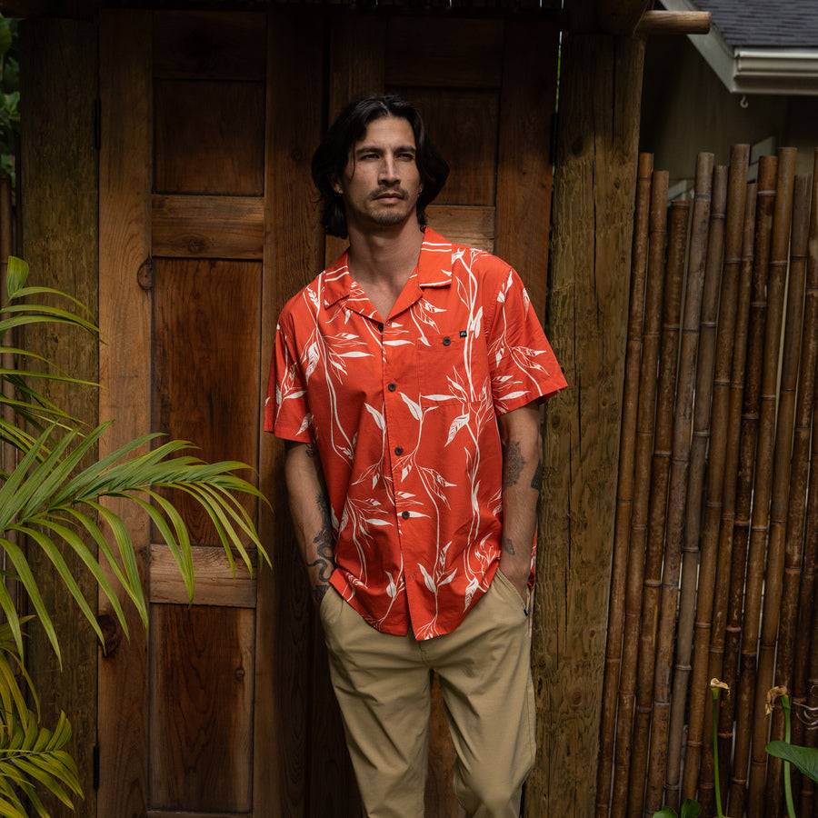 Man wearing a red Kaden Camp Shirt with white tropical print, standing relaxed by a wooden door outdoors.