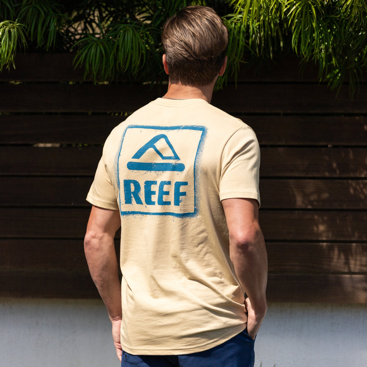 Man wearing beige Xavier Tee with blue REEF logo on back, standing outdoors facing away, beach-inspired comfort apparel.