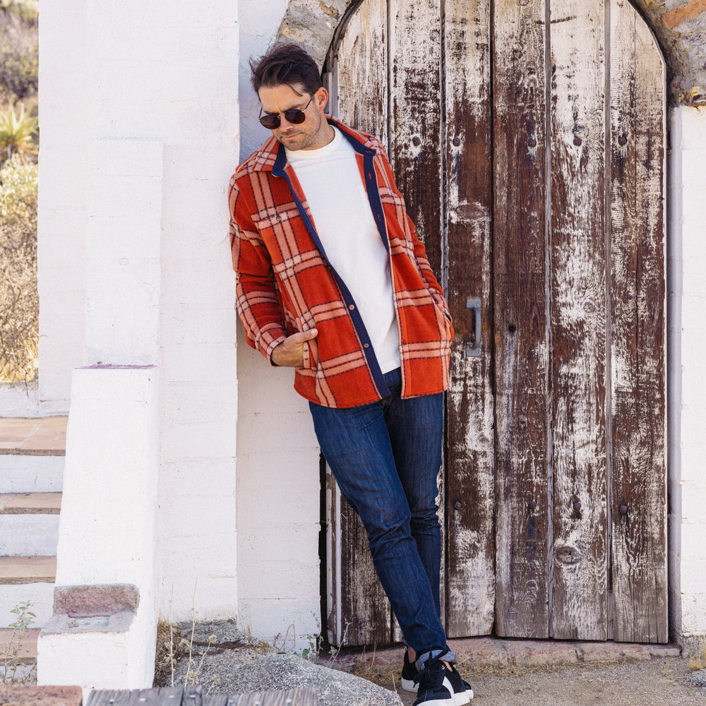 Man wearing Karter Shirt Jacket in cinnabar plaid leaning by rustic wooden door, styled for a beach-inspired look