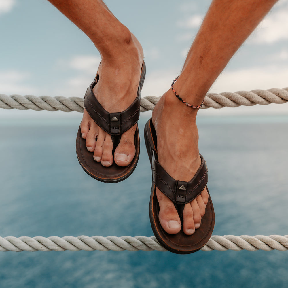 Leather Offshore men's sandals in dark brown, front facing close-up showing padded straps and cushioned footbed by the ocean.