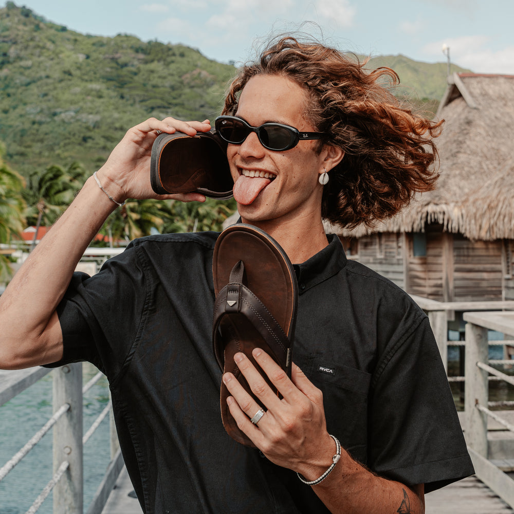 Man holding a pair of Leather Offshore sandals, one facing forward and the other showing the outsole, beach background.