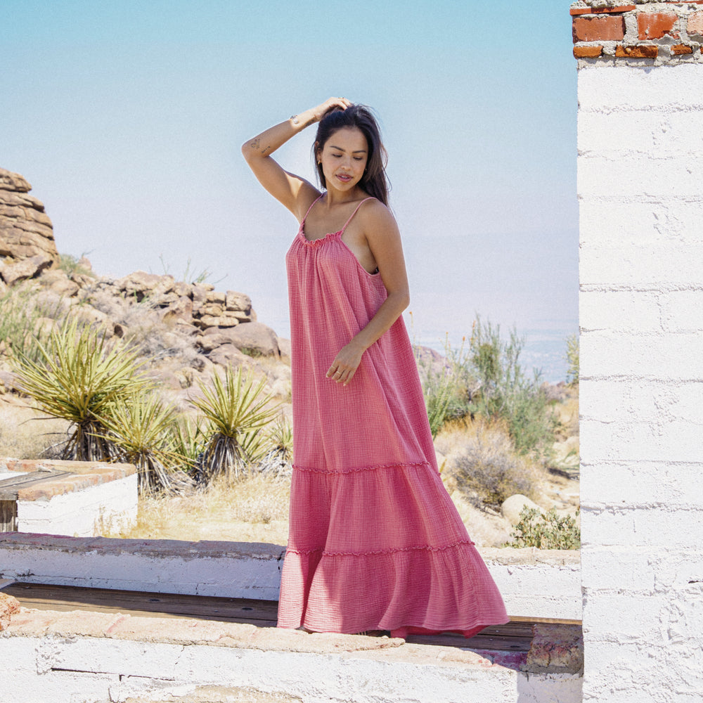 Woman wearing a mauve Lainey Maxi Dress standing outdoors by rustic wall in sunny desert setting with light breeze