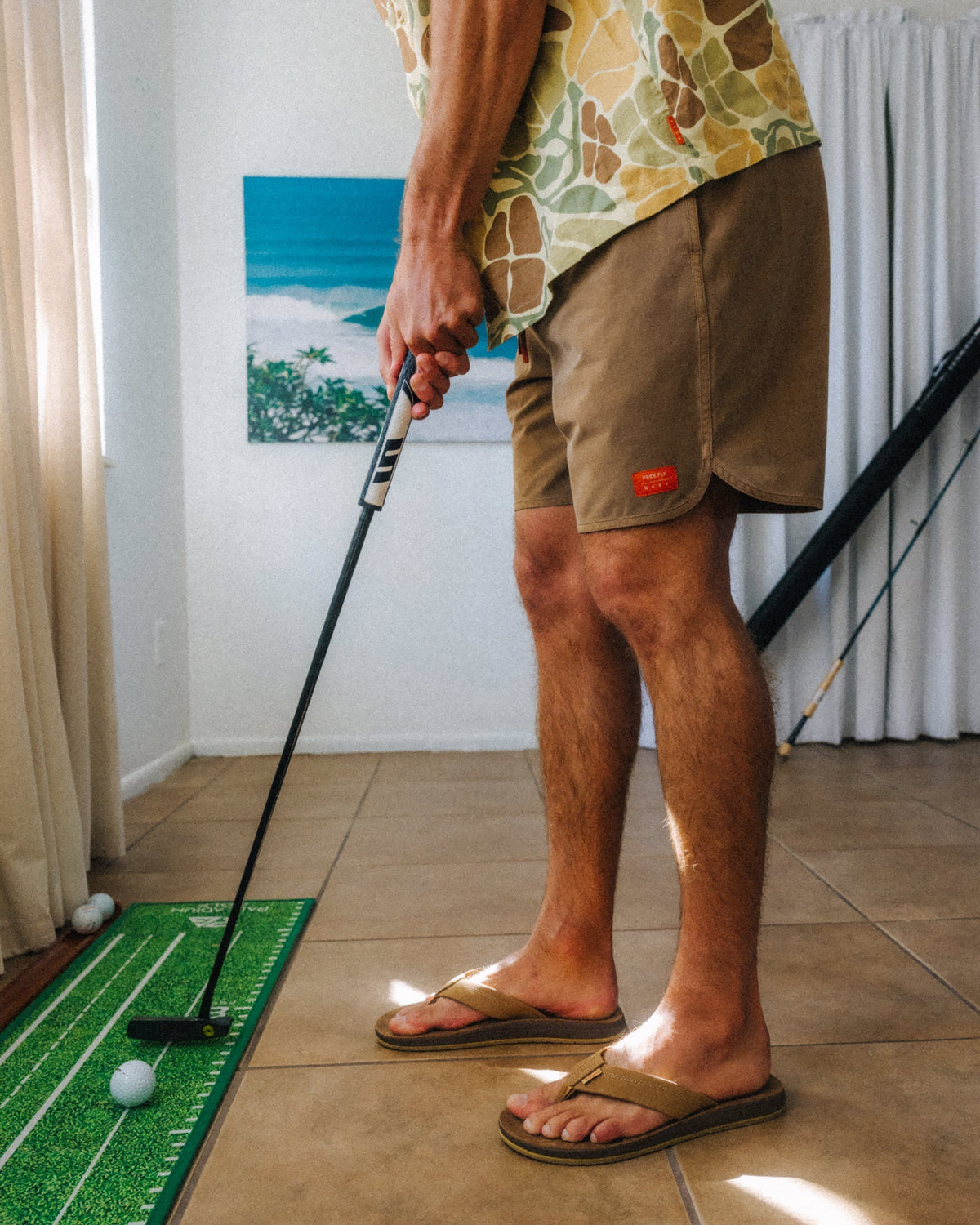 Man wearing Men's Reverb Short Unlined – 7" - Coconut with tan sandals facing right indoors playing mini golf on green mat