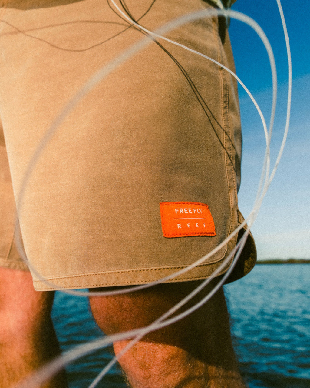 Close-up of Men's Reverb Short Unlined – 7" - Coconut in tan with visible orange brand patch, beach background, facing right