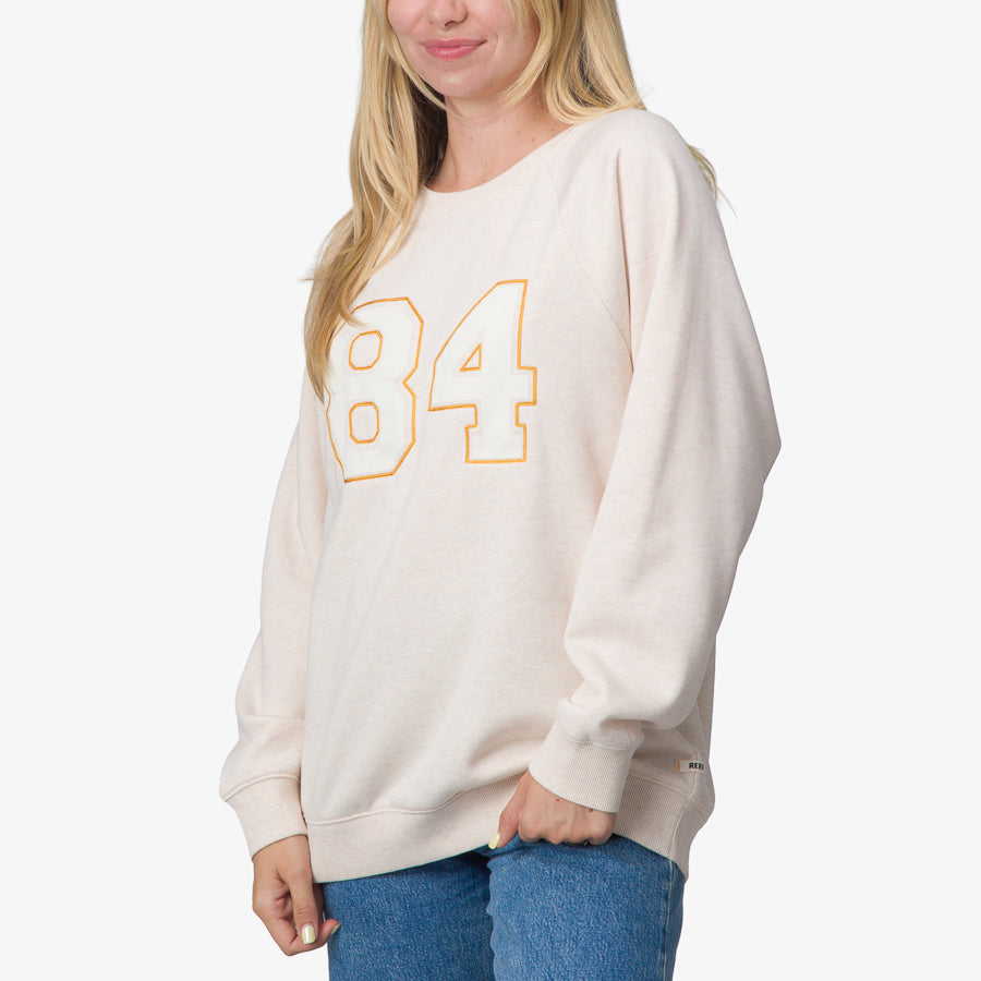 Woman wearing an oatmeal oversized raglan fleece with number 84 graphic, shown front and angled left, comfort-focused apparel for beach living