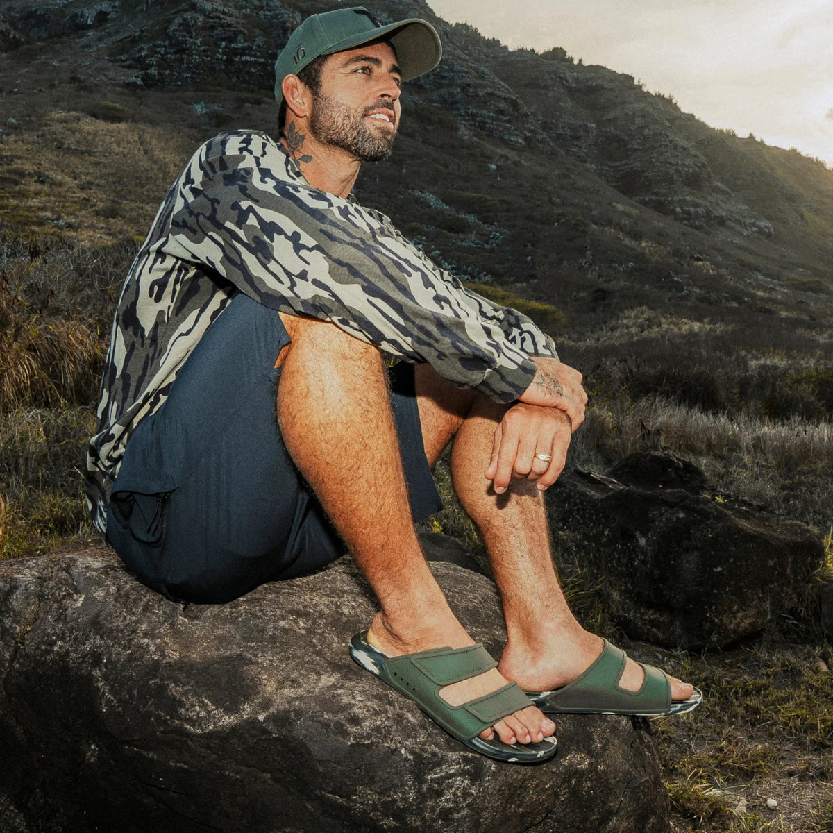 Oasis Adapt sandals facing right on man sitting on rock in camo apparel, showcasing comfort and water-friendly design.