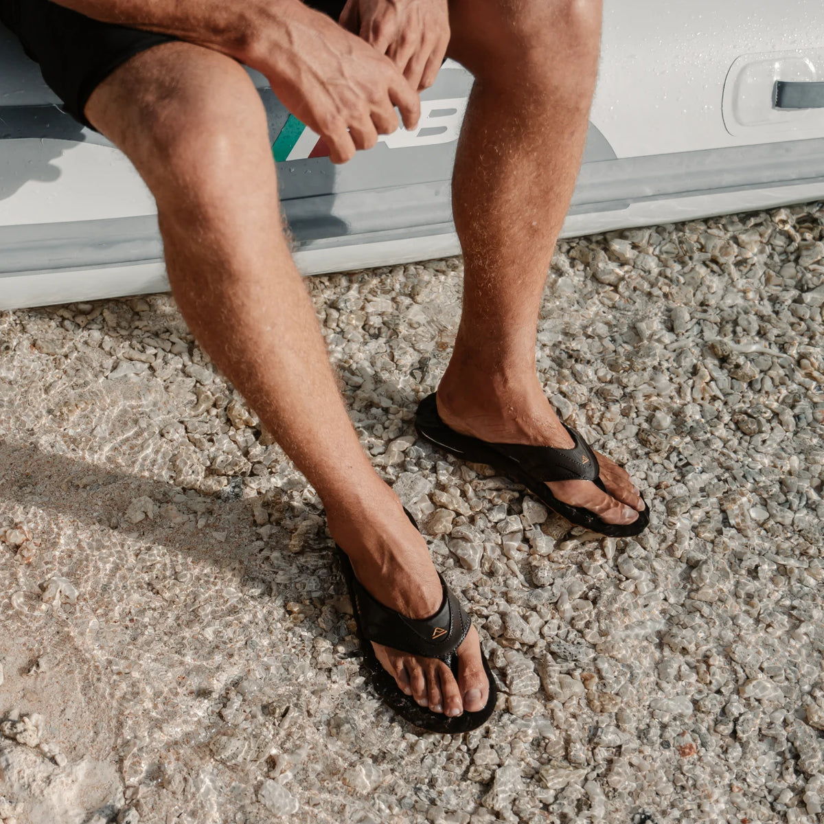 Fanning flip flops facing forward on feet, showing black synthetic upper and cushioned footbed with arch support by the beach.