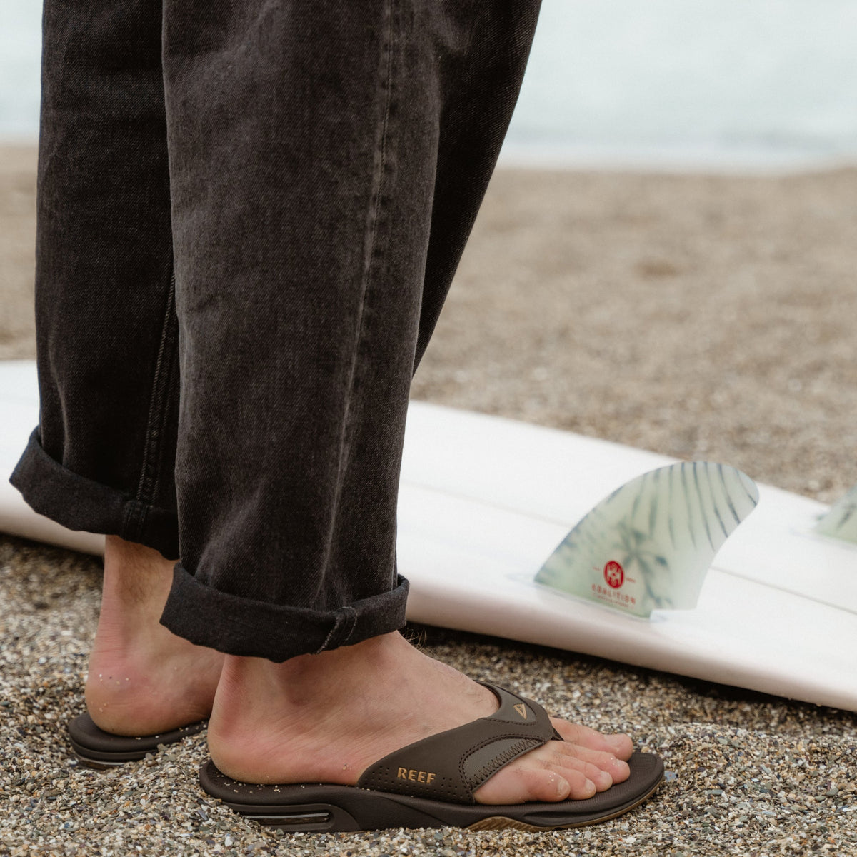 Fanning sandals facing right on a man’s feet with anatomical arch support, soft cushioning, on a beach near a surfboard fin.