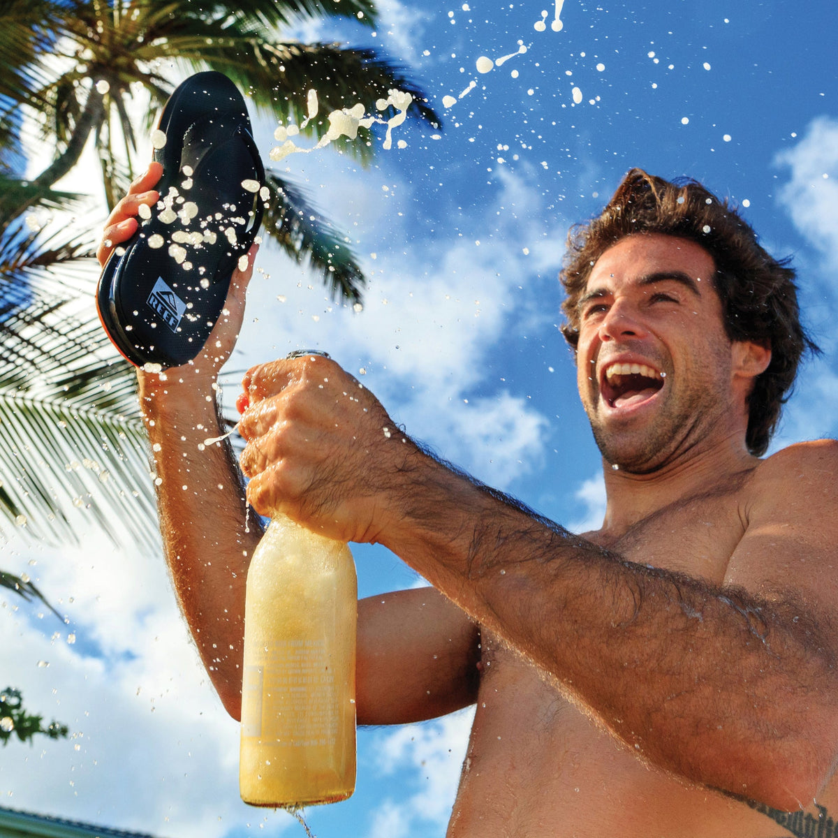 Man holding Fanning sandal facing up in beach setting with palm trees, showing sole and water splashing around him