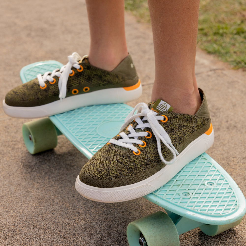 Close-up of Kids Neptune sneaker in olive green facing right with white laces on blue skateboard, showcasing comfort and durability.
