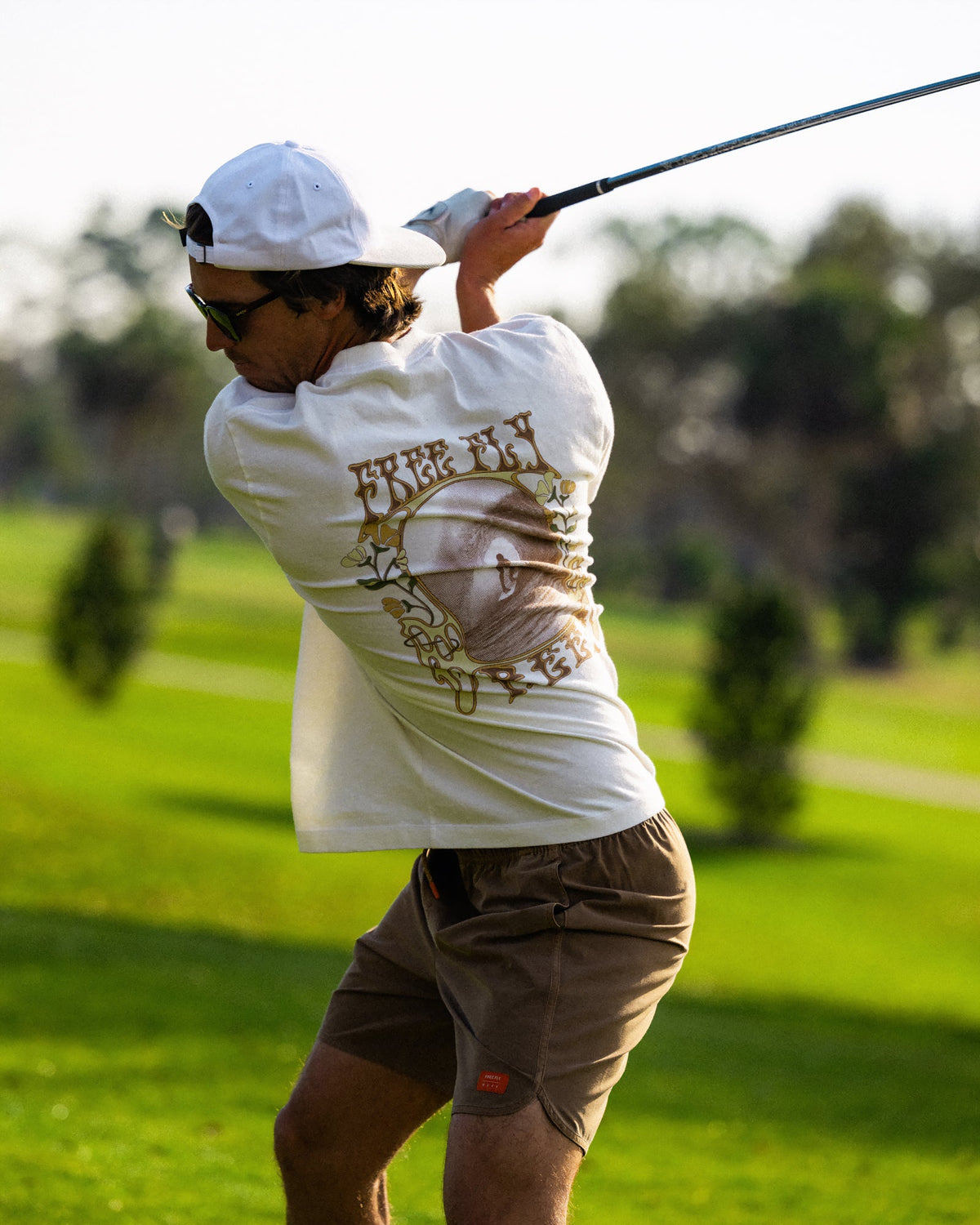 Man swinging golf club wearing High-Stoke Relaxed Tee - Bone with brown shorts, casual beach-inspired athletic look outdoors