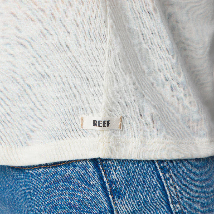 Close-up of the bottom hem of the Ringer Tee in marshmallow white, showing REEF logo tag above blue denim jeans.