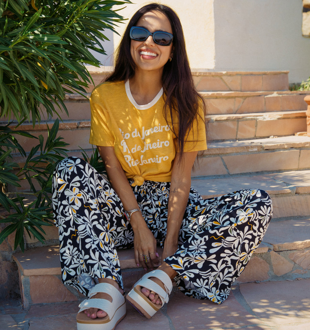 Woman wearing Ringer Tee in honey gold with floral pants sitting outdoors, showcasing beach-inspired comfort apparel.