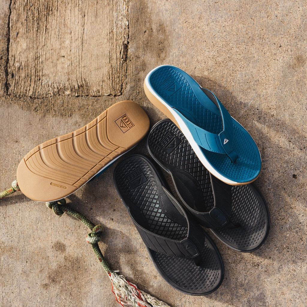Pair of Deckhand flip flops for men, one blue facing up showing sole detail, two black facing down with arch support design.