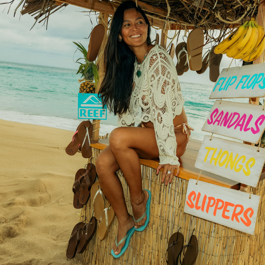Woman wearing Solaye teal sandals facing slightly left, sitting on beach hut counter with surf-inspired footwear displayed nearby.