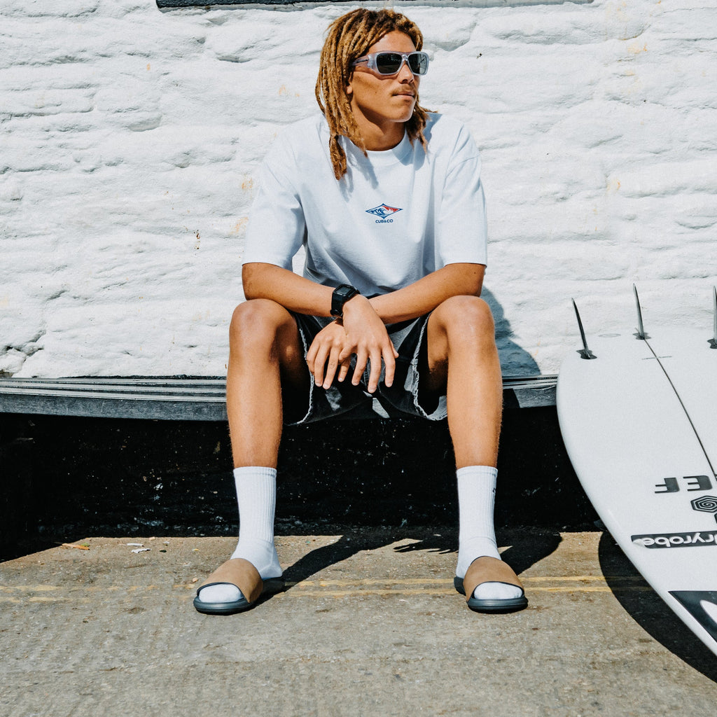 Tailslide men's tan slides with toes facing the camera on a seated model beside a surfboard