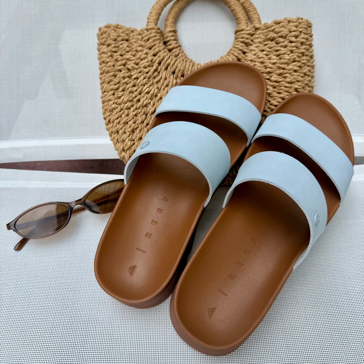 Vista Luxe Thea sandals facing upwards with blue straps, brown footbed, beside woven bag and sunglasses.
